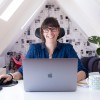 A woman sits smiling at her desk with an open laptop and wearing a hat.