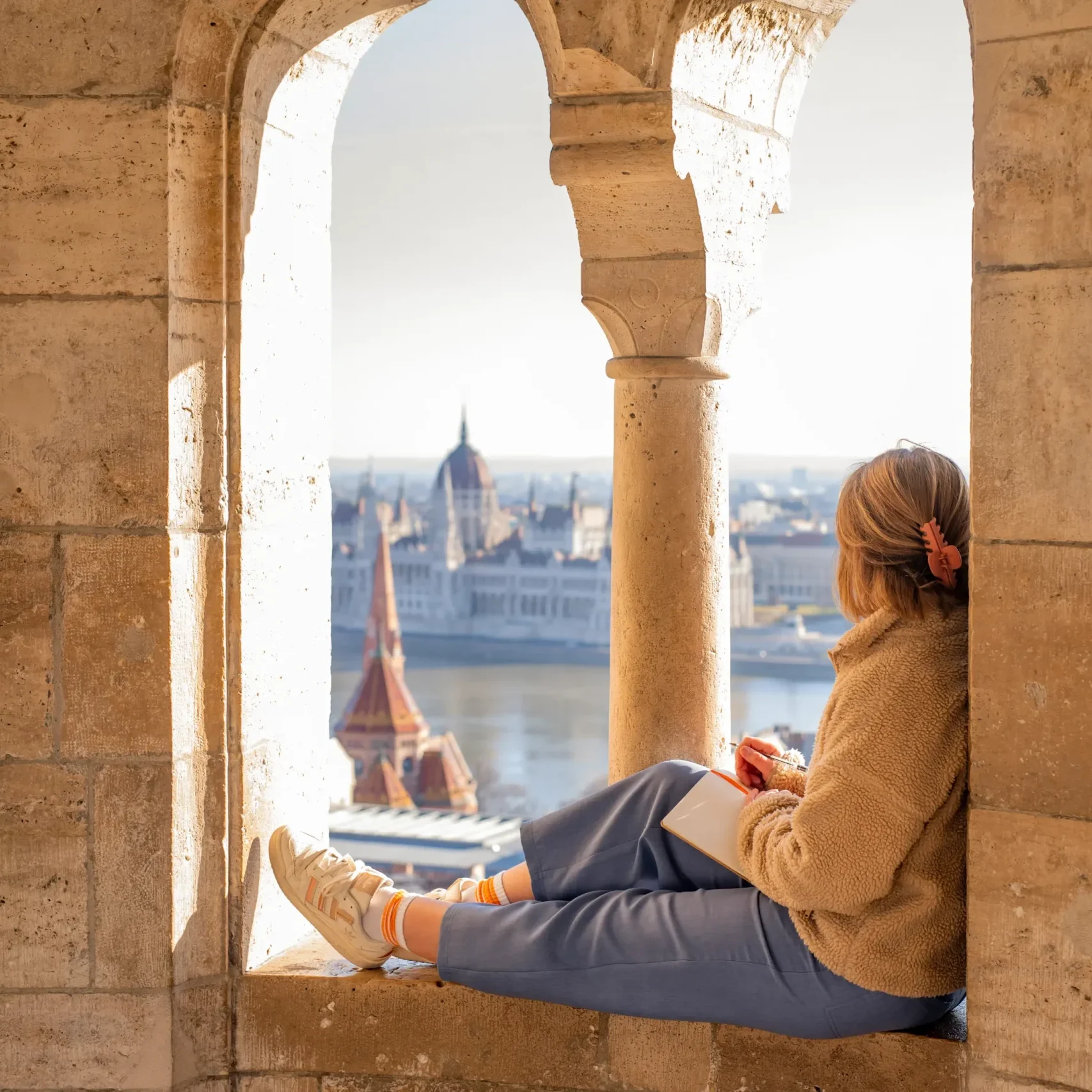 A woman writes about the business of storytelling in a stone archway at Fisherman’s Bastion, looking out at Budapest.