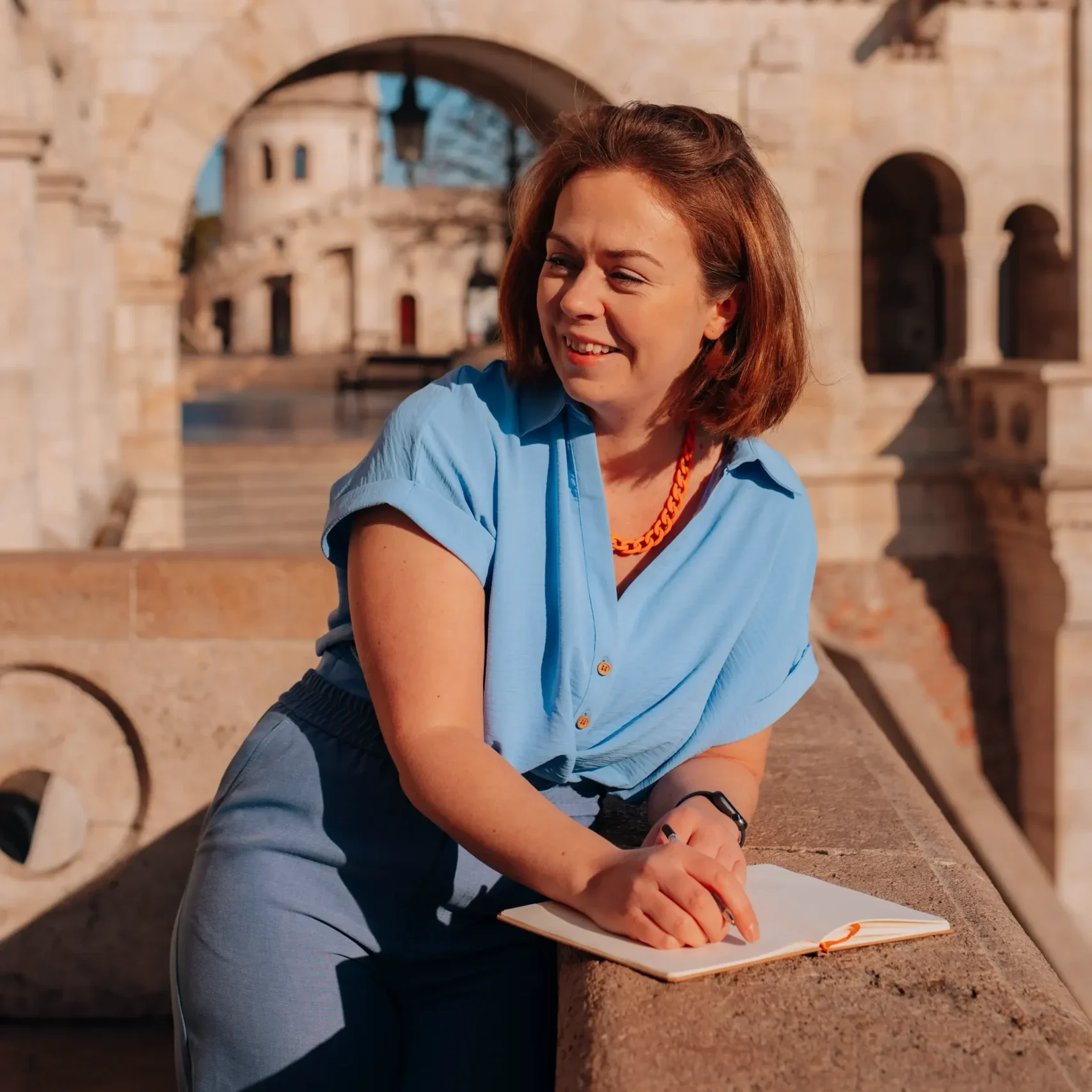 A creative copywriter smiling while writing in a notebook, standing at Fisherman’s Bastion in Budapest, Hungary.