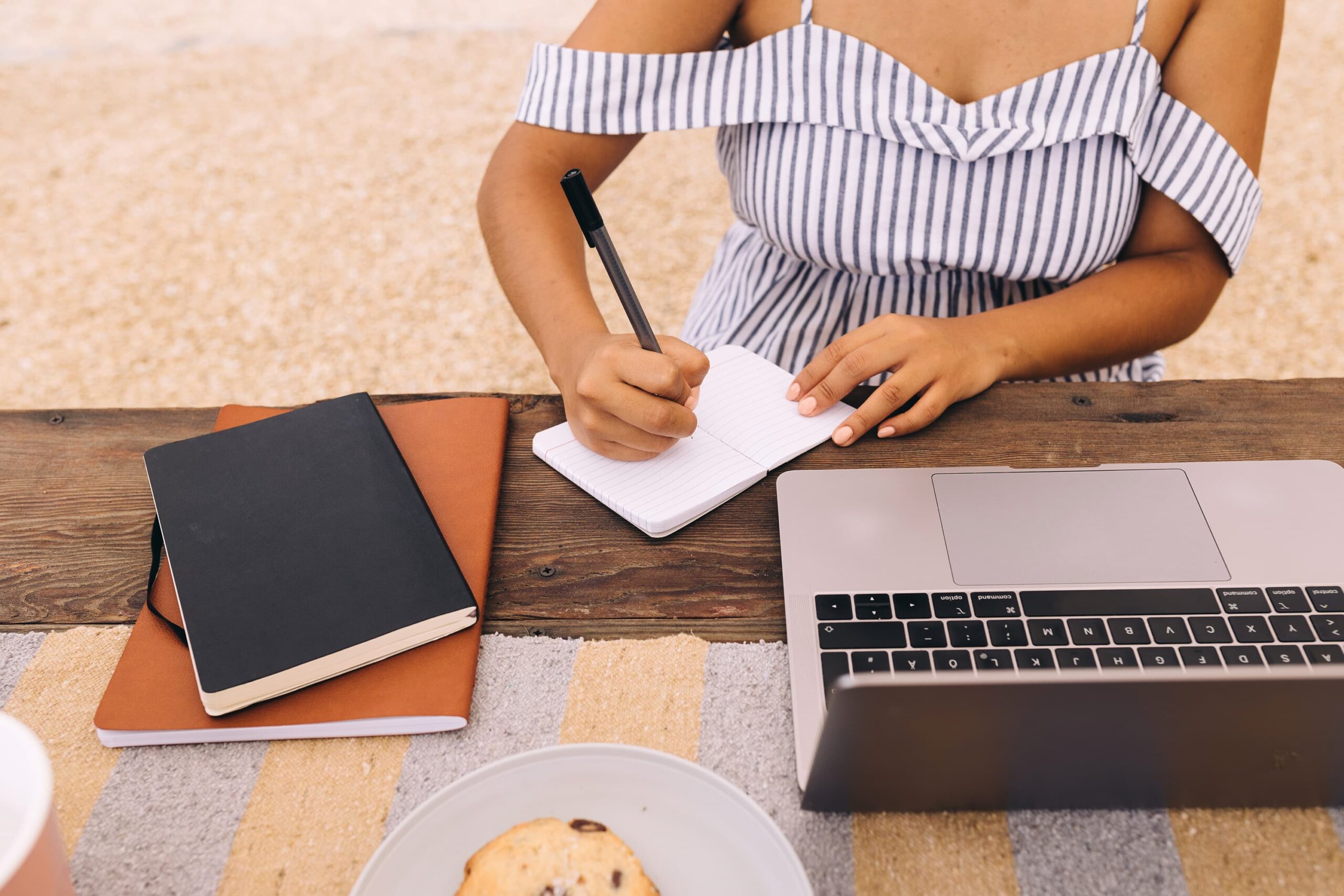 A woman in a striped summer dress writing in a notebook at a wooden table with a laptop, showcasing the creative process behind brand storytelling.
