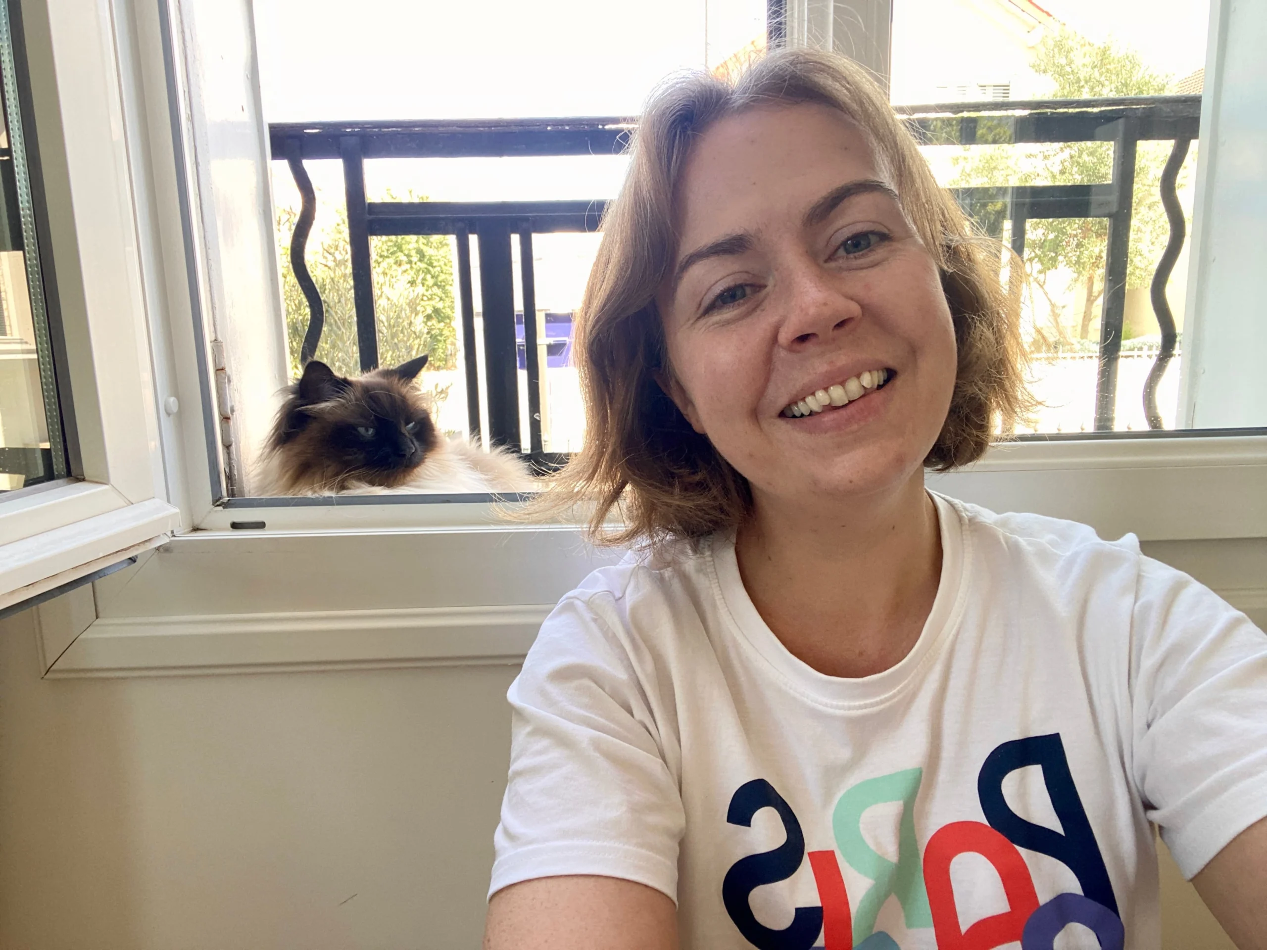 Woman in a white t-shirt smiling at the camera with a cat sitting on a windowsill in the background.