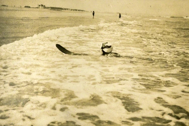Vintage sepia photograph of Agatha Christie surfing in ocean waves with figures on beach