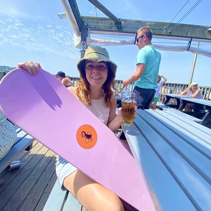 Woman holding wooden bellyboard promoting sustainable surf culture at outdoor beach bar on sunny day