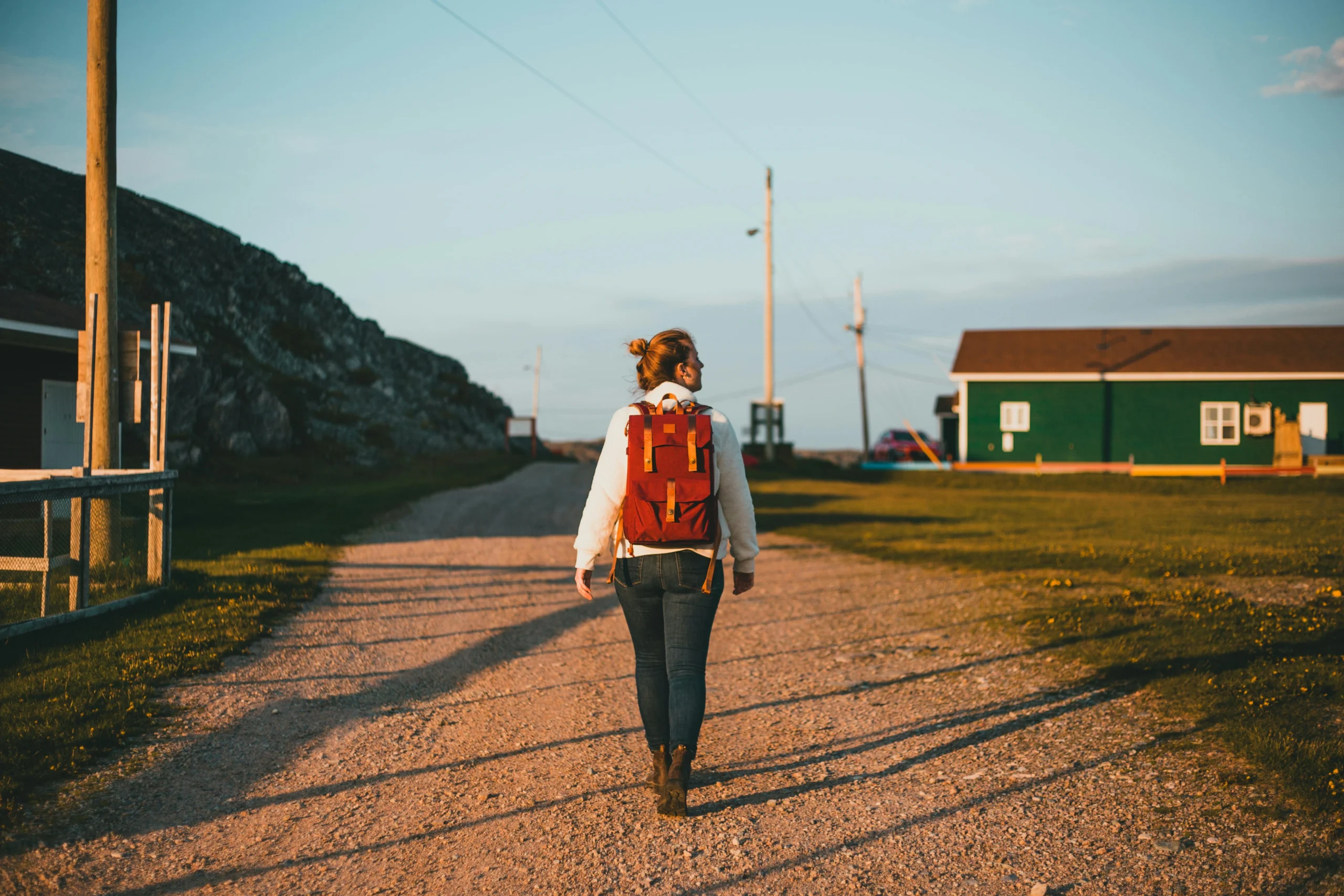 Solo female traveller walking down a quiet rural road with a red backpack at sunset.