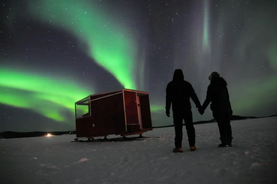 Two people standing beside a mobile cabin on frozen Lake Inari beneath the northern lights.