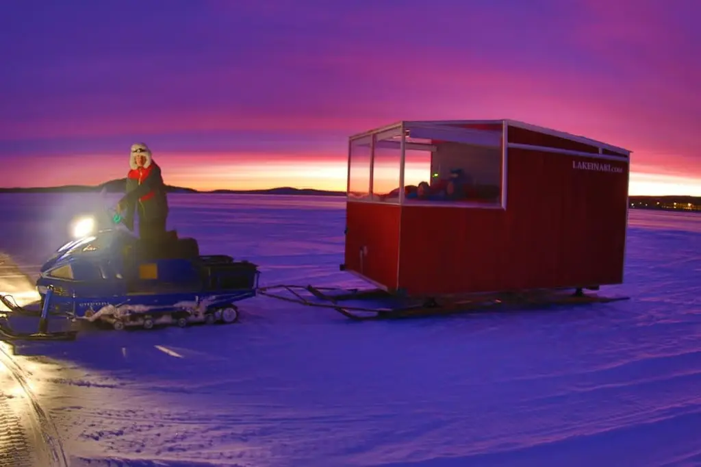 A mobile cabin being towed across the frozen lake by a snowmobile at sunset.
