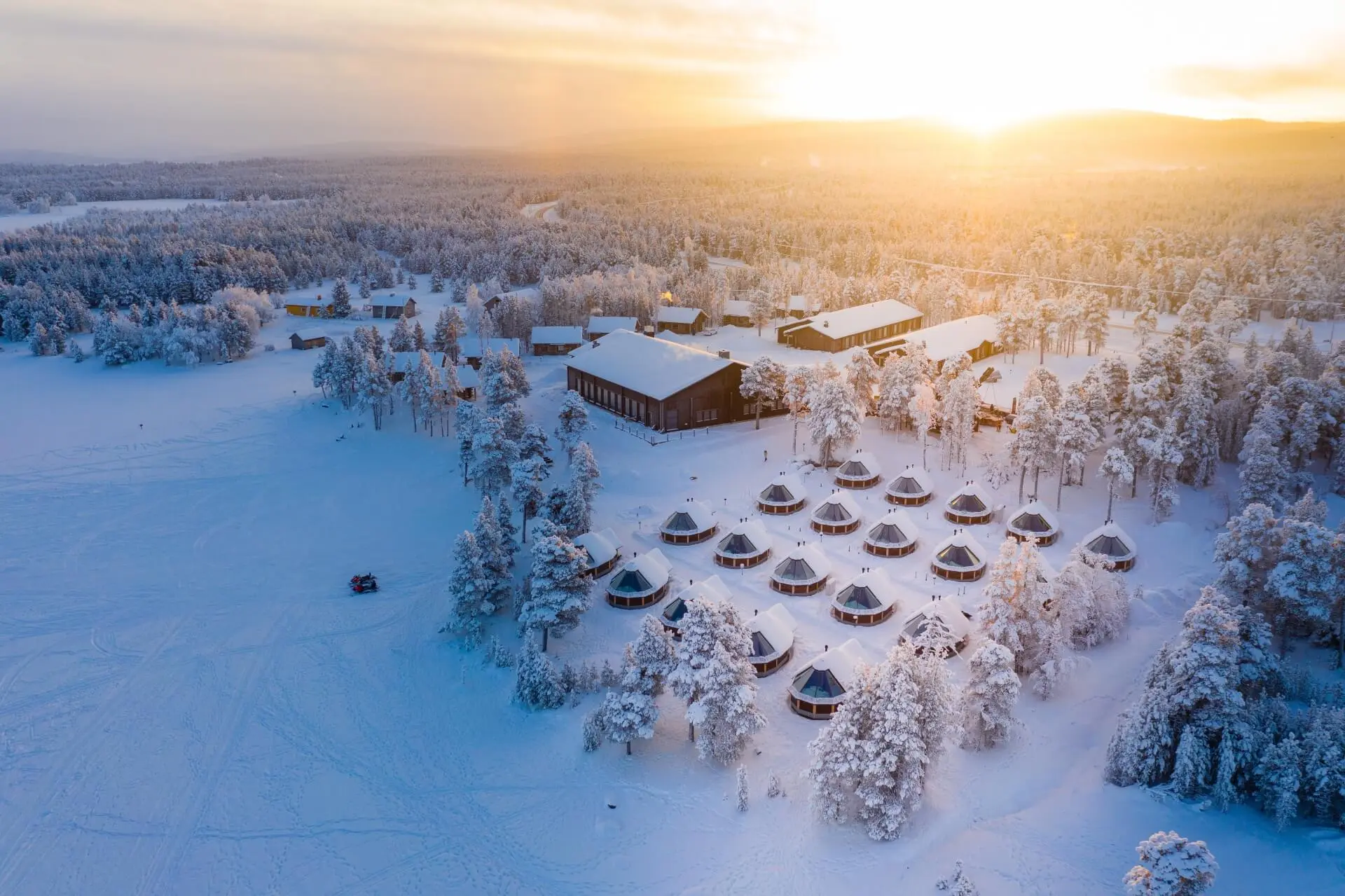Aerial view of Wilderness Hotel Inari surrounded by snow-covered trees and cabins, with the winter sun low on the horizon.