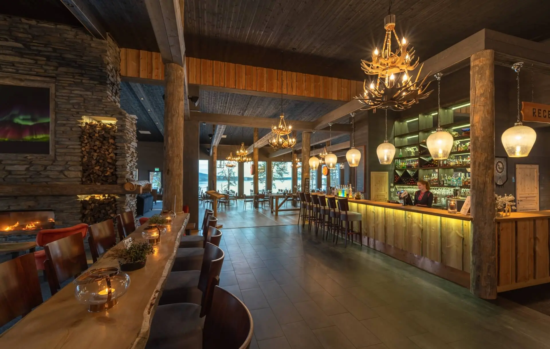 Rustic restaurant and bar inside Wilderness Hotel Inari, with wooden interiors, a large stone fireplace and warm lighting.