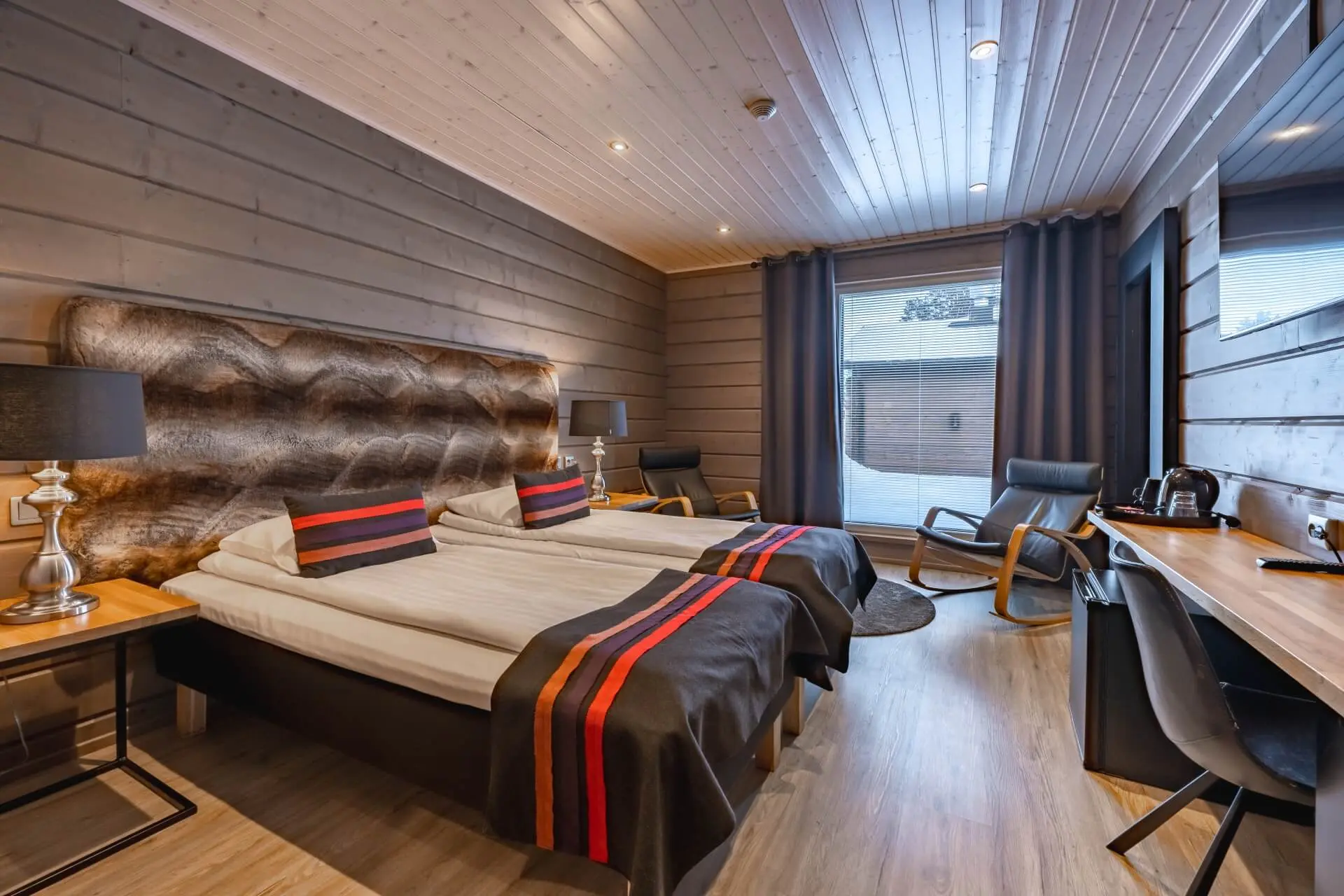 Cosy bedroom at Wilderness Hotel Inari, with a large bed, wooden walls, a desk and a window overlooking the snowy landscape.